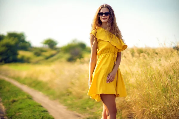 Hermosa chica rizada camina en el campo en verano mientras usa gafas de sol y vestido amarillo. Estilo de vida —  Fotos de Stock