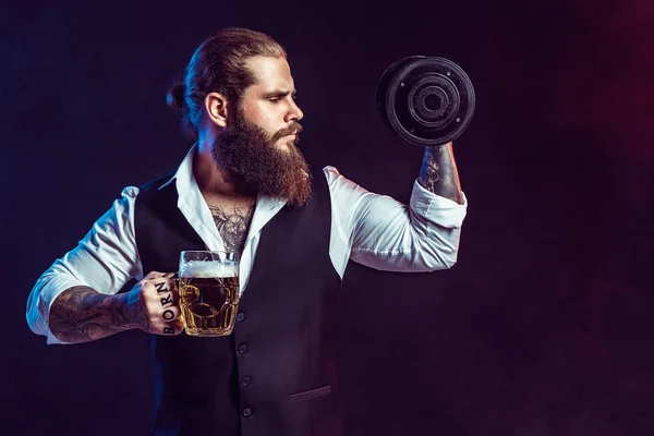 Bearded man holds dumbbell and mug of beer on dark background while choices alcohol or sport — Zdjęcie stockowe