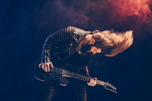 黒の背景に長い髪とひげを生やしたロックギタープレーヤーの感情的な肖像画 — ストック写真