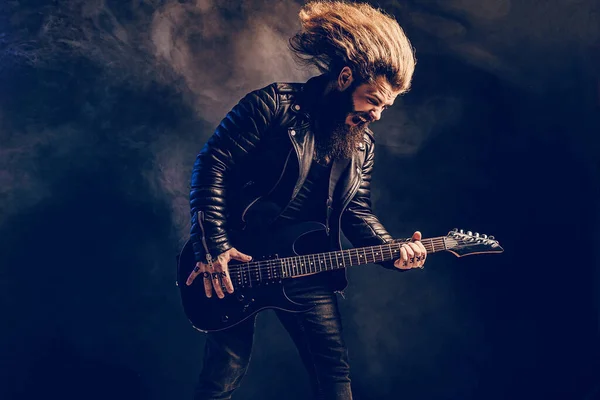 長い髪とひげを持つ感情的な男ロックギタープレーヤーは煙の背景で再生されます。スタジオショット — ストック写真