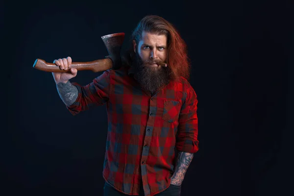 man with cigar holds old axe. Bearded lumberjack. Serious man in checkered shirt with long hair