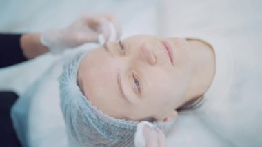 Dermatolog doktor, kozmetik prosedürü için hastanın yüzünü dezenfekte bileşimiyle siliyor.