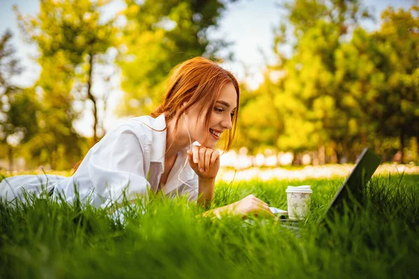 Молода красива руда щаслива дівчина використовує ноутбук ПК і навушники, лежачи на траві в парку — стокове фото