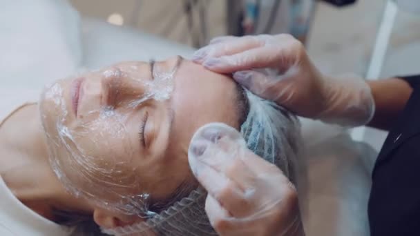 В клинике красоты косметолог очищает кожу лица молодой клиентки ватным тампоном — стоковое видео