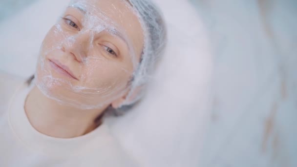 Natuurlijke mooie jonge vrouw met cosmetische masker op haar gezicht ontspannen in schoonheidssalon. Huidverzorging — Stockvideo