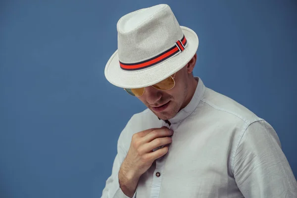 Studio portrét stylového muže, který pózuje ve slunečních brýlích s elegantním bílým kloboukem na modrém pozadí — Stock fotografie