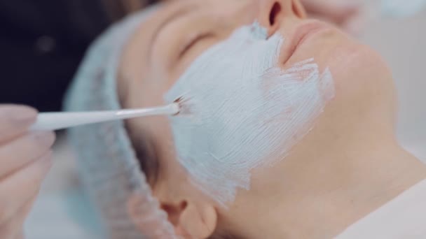 Kosmetikerin trägt Gesichtsmaske mit Pinsel auf junge Patientin in Klinik auf. Kosmetische Hautpflege. — Stockvideo