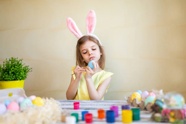 Skoncentrowana dziewczynka w króliczych uszach malująca jajka wielkanocne w domu przygotowuje się do Wielkanocy. Wesołych Świąt — Zdjęcie stockowe