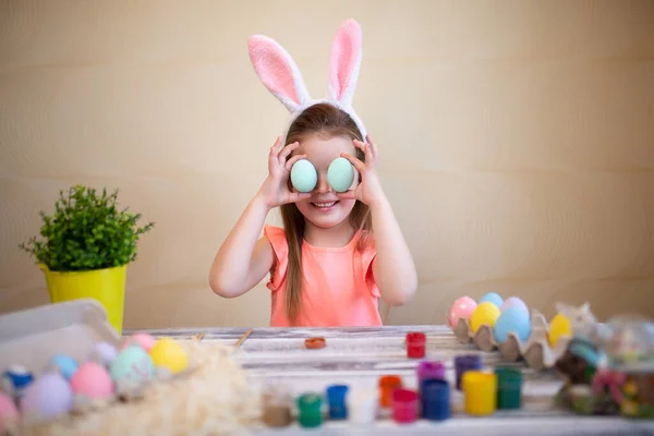 Szczęśliwa dziewczynka trzyma wielkanocne jajka w pobliżu oczu przygotowując się do Wielkanocy nosząc królicze uszy. — Zdjęcie stockowe