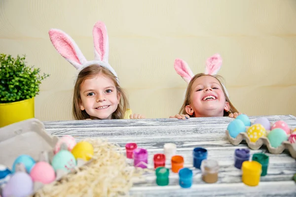Szczęśliwe dwie małe dziewczynki w króliczych uszach wyjrzały zza stołu z kolorowymi pisankami. — Zdjęcie stockowe