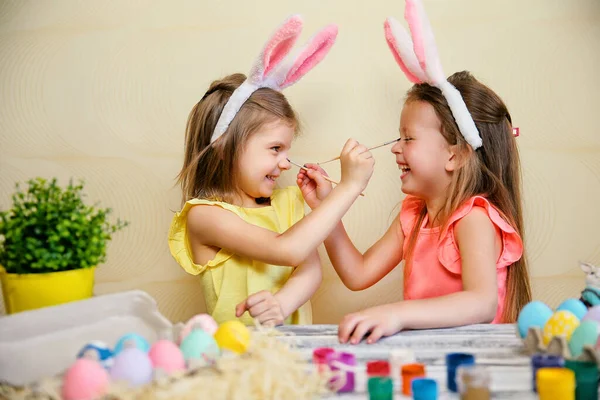 Dwie małe dziewczynki w króliczych uszach malują wielkanocne jajka i obmacują się nawzajem. Przygotowania do Wielkanocy — Zdjęcie stockowe