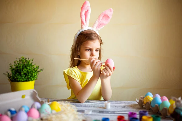 Cute girl in bunnys uszy maluje pisanki w domu siedząc przy stole. Przygotowania do Wielkanocy — Zdjęcie stockowe