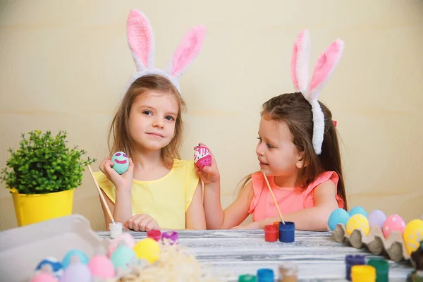 Dwie dziewczynki w króliczych uszach biją wielkanocne jajka siedząc przy stole. Szczęśliwej koncepcji Wielkanoc — Zdjęcie stockowe