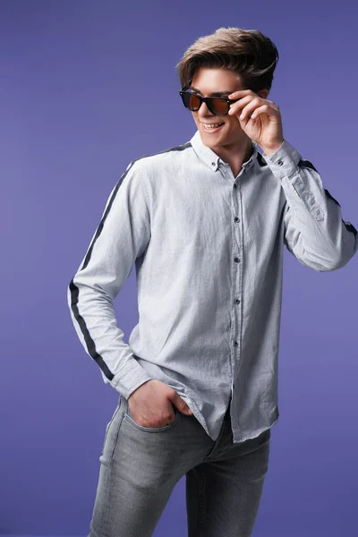 Glimlachende hipster in stijlvolle zonnebril houdt arm in zak, geïsoleerd op paarse achtergrond — Stockfoto