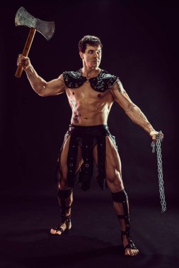 Siyah arka planda poz verirken balta ve zincir tutan güçlü bir savaşçı..