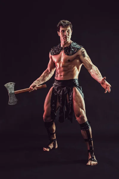Teljes méretű izmos gladiátor, aki fejszét tart, és félrenéz, miközben pózol a füst háttér. — Stock Fotó