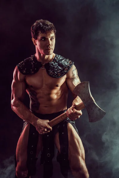 Porträt eines sexy jungen Kriegers, der die Axt in der Hand hält und wegschaut, während er im Hintergrund posiert. — Stockfoto