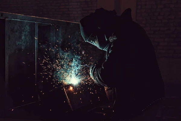 Boční pohled na svářečku při práci. Dělník svařuje kovové části. Svařování, jiskry, plamen, kouř. — Stock fotografie