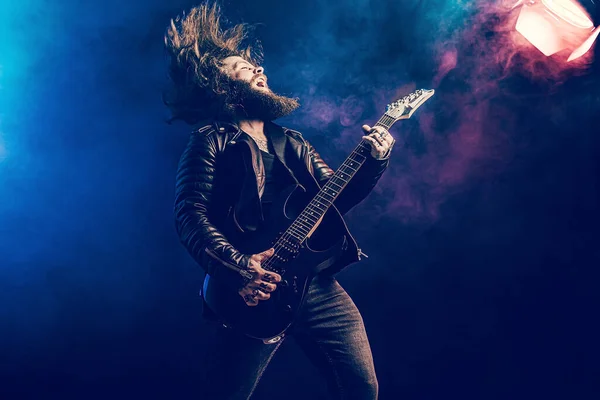 Emotional man rock guitar player with long hair and beard plays on the smoke background. Studio shot — Stock Photo, Image
