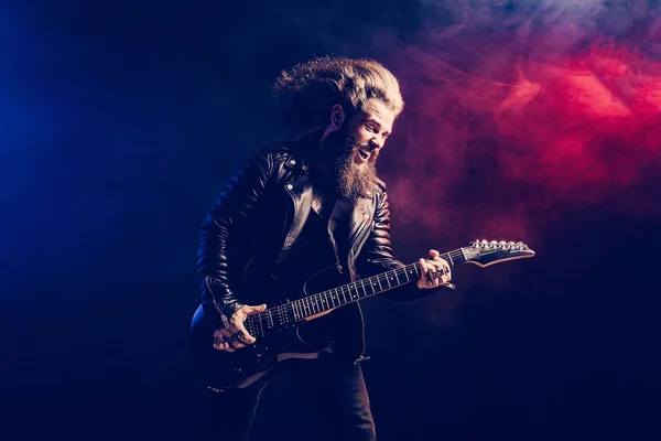 煙の背景に長い髪とひげを生やした表現力豊かな男性ロックギタープレーヤー。スタジオショット — ストック写真