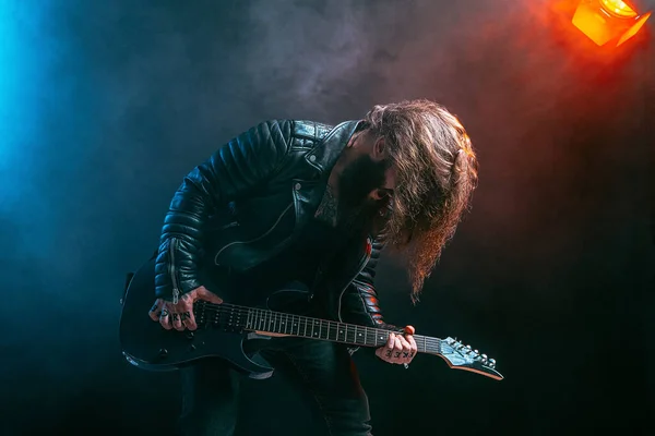 Érzelmes férfi rock gitáros hosszú hajjal és szakállal játszik a füst háttérben. Stúdió felvétel — Stock Fotó