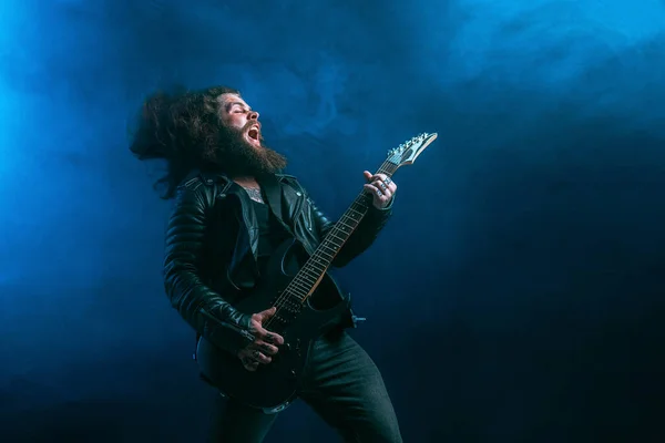 Emotional man rock guitar player with long hair and beard plays on the smoke background. Studio shot — Stock Photo, Image