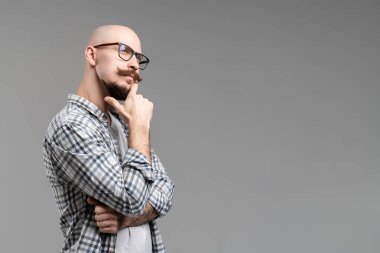 Düşünceli, sakallı, gözlüklü adam, gri arka planda izole bir şekilde çenesini yumrukluyor.