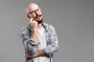 Gri arka planda izole bir şekilde çenesine yumruk atan gözlüklü şüpheli sakallı adamın portresi.