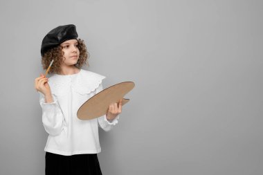 Küçük kız ressam gri stüdyo arka planında boya fırçası ve palet tutuyor. Çocuk hobisi kavramı