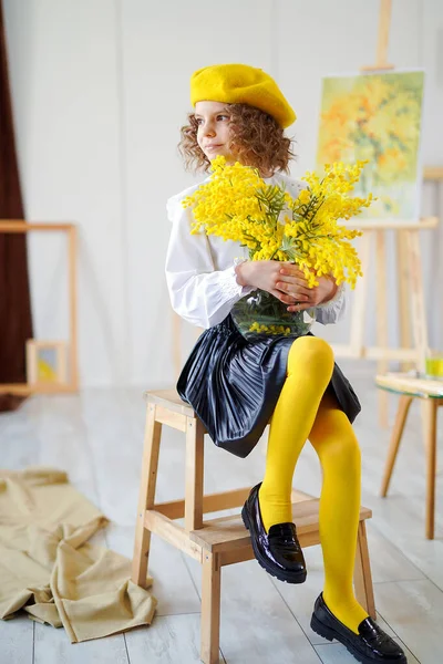 Comprimento total da menina encaracolado em boina amarela que posa em estúdio, enquanto segurando flores amarelas — Fotografia de Stock