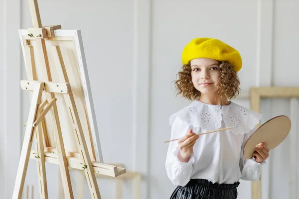 Retrato de menina artista em boina amarela que pinta imagem em estúdio, enquanto segura palete — Fotografia de Stock
