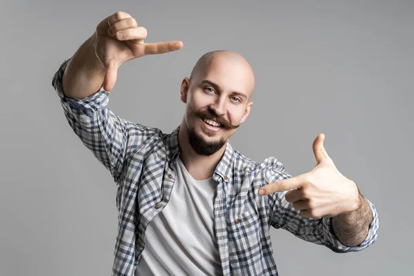 Χαμογελώντας φαλακρός γενειοφόρος άνθρωπος κάνει πλαίσιο με τα δάχτυλα μπροστά από το πρόσωπο πάνω από γκρι φόντο — Φωτογραφία Αρχείου