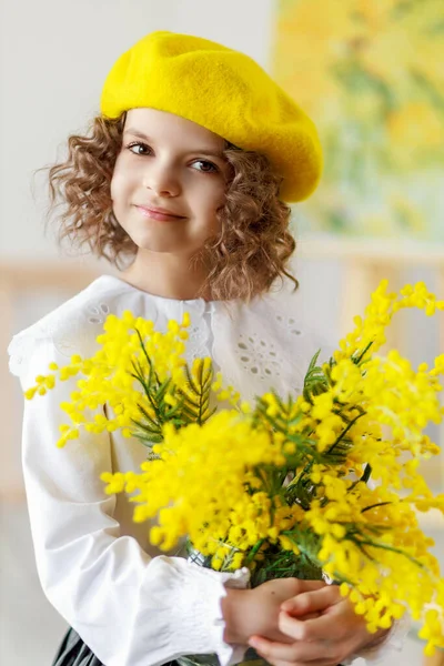 Retrato de una niña rizada linda en boina amarilla y medias que posa en el estudio mientras sostiene flores amarillas — Foto de Stock