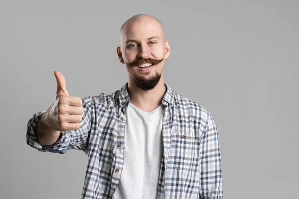 Young happy bald bearded man in checkered shirt gestures thumb up isolated on gray background — Stock Photo, Image