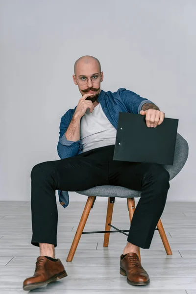 Хіпстер бородатий чоловік з вусами в окулярах, який сидить на стільці в студії ізольований сірий фон тримає кишеню, серйозно виглядаючи — стокове фото