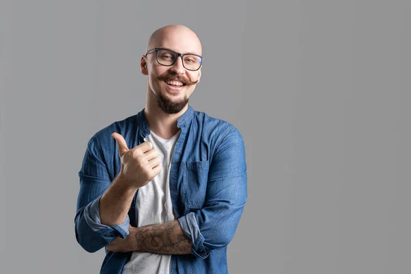 Portrait of a smiling balded bearded with mustache man in glasses showing thumb up over gray background — Stockfoto