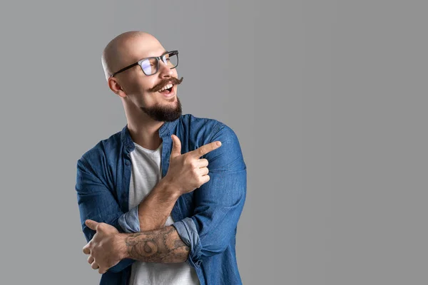 Young man with moustache smiling with happy face looking and pointing to the side with index finger — Stockfoto