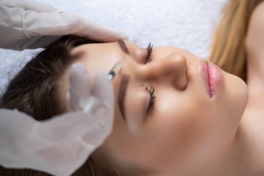 Çekici genç bir kadın canlandırıcı yüz enjeksiyonu yaptırıyor. O, klinikte sakince oturuyor. Uzman güzellik uzmanı hyaluronik asitle kadın kırışıklıklarını dolduruyor.. 