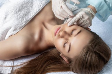 Çekici genç bir kadın canlandırıcı yüz enjeksiyonu yaptırıyor. O, klinikte sakince oturuyor. Uzman güzellik uzmanı hyaluronik asitle kadın kırışıklıklarını dolduruyor.. 