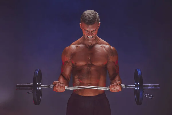 Brutal sweaty strong young man athlete with naked upper body standing doing workout with barbell and showing strong pumped up biceps over smoky background. Sport men body concept.