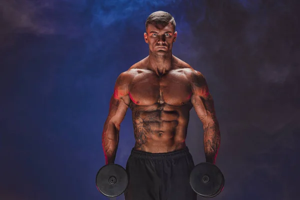 Brutal sweaty strong young man athlete with naked upper body standing doing workout with dumbbels and showing strong pumped up biceps over smoky background. Sport men body concept.