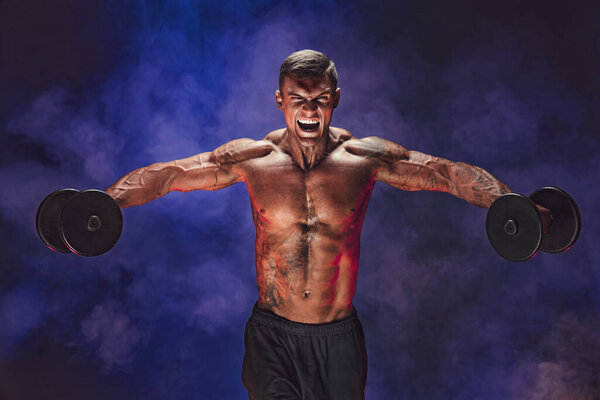 Brutal sweaty strong young man athlete with naked upper body standing doing workout with dumbbels and showing strong pumped up biceps over smoky background. Sport men body concept. 