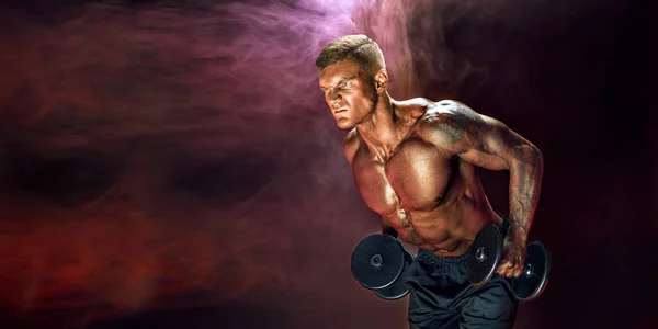 Brutal sweaty strong young man athlete with naked upper body standing doing workout with dumbbels and showing strong pumped up biceps over smoky background. Sport men body concept.