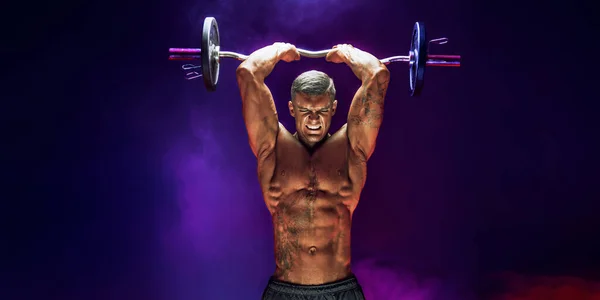 Brutal sweaty strong young man athlete with naked upper body standing doing workout with barbell and showing strong pumped up biceps over smoky background. Sport men body concept.