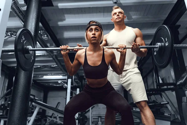 A young male personal trainer helps a woman to stimulate the muscles while she does squats. Fitness modern gym concept.