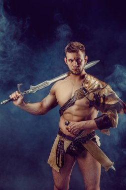 Portrait of handsome muscular gladiator with sword. Isolated. Studio shot. Dark background. 