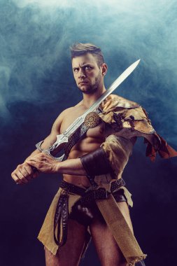 Portrait of handsome muscular gladiator with sword. Isolated. Studio shot. Dark background. 