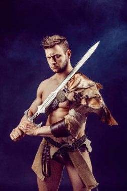 Portrait of handsome muscular gladiator with sword. Isolated. Studio shot. Dark background. 