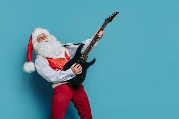 Emotional Santa Claus plays on electric guitar on blue studio background. Christmas music. New Year party