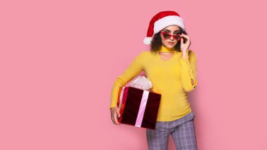 Young smiling happy cheerful curly girl in Santas hat holds red giftbox while stands on pink background. Sopping.
